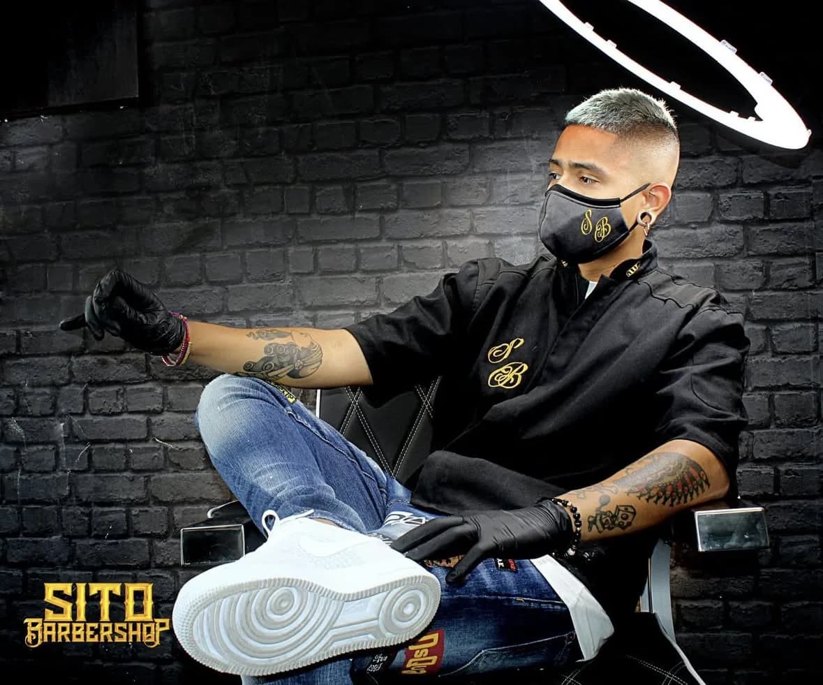Sito, el barbero, sentado en su sillón frente a una pared de ladrillo negro, con guantes, mascarilla bordada SB y un aro de luz encima